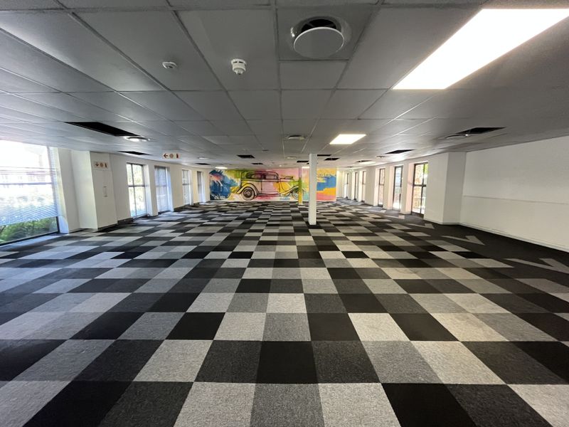 A large, open-plan office space featuring a checkered carpet tile floor, suspended ceiling with recessed lighting, and large windows providing natural light.