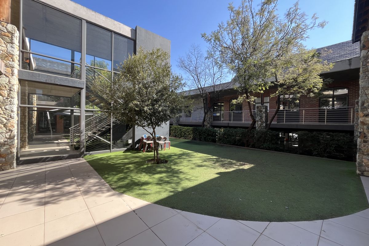 A modern office courtyard featuring a glass-walled building, stone accents, a steel staircase, and a well-maintained artificial lawn area.