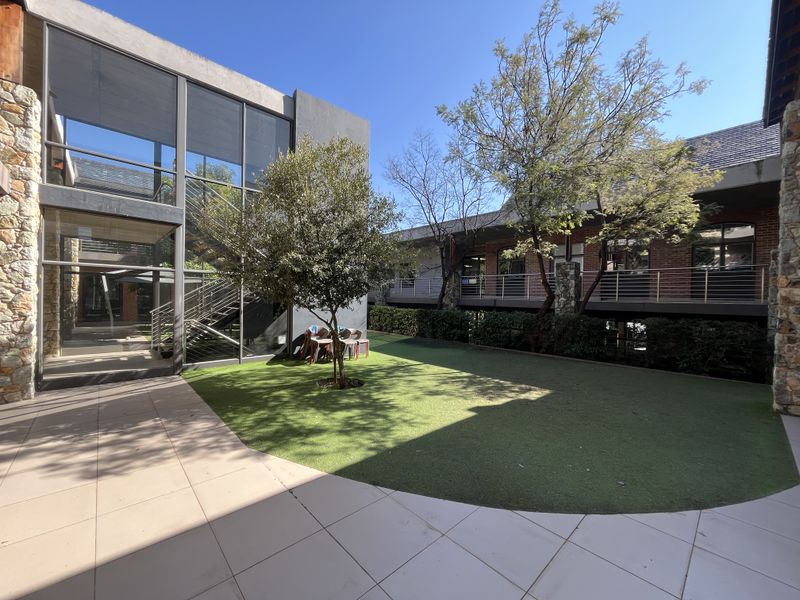A modern office courtyard featuring a glass-walled building, stone accents, a steel staircase, and a well-maintained artificial lawn area.
