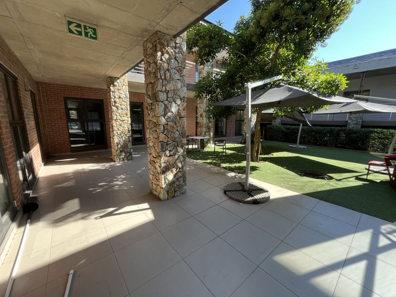 A covered outdoor patio area featuring stone-clad columns, exposed concrete ceilings, and tiled flooring, adjacent to a landscaped garden space.
