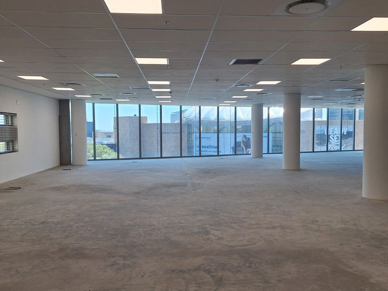 A spacious, open-plan office area featuring large floor-to-ceiling windows, a suspended ceiling with recessed lighting, and unfinished concrete flooring.