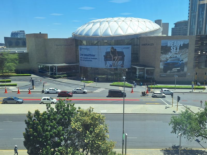 An elevated view overlooking the Sandton City shopping centre entrance, featuring its iconic dome roof and surrounding urban street infrastructure.
