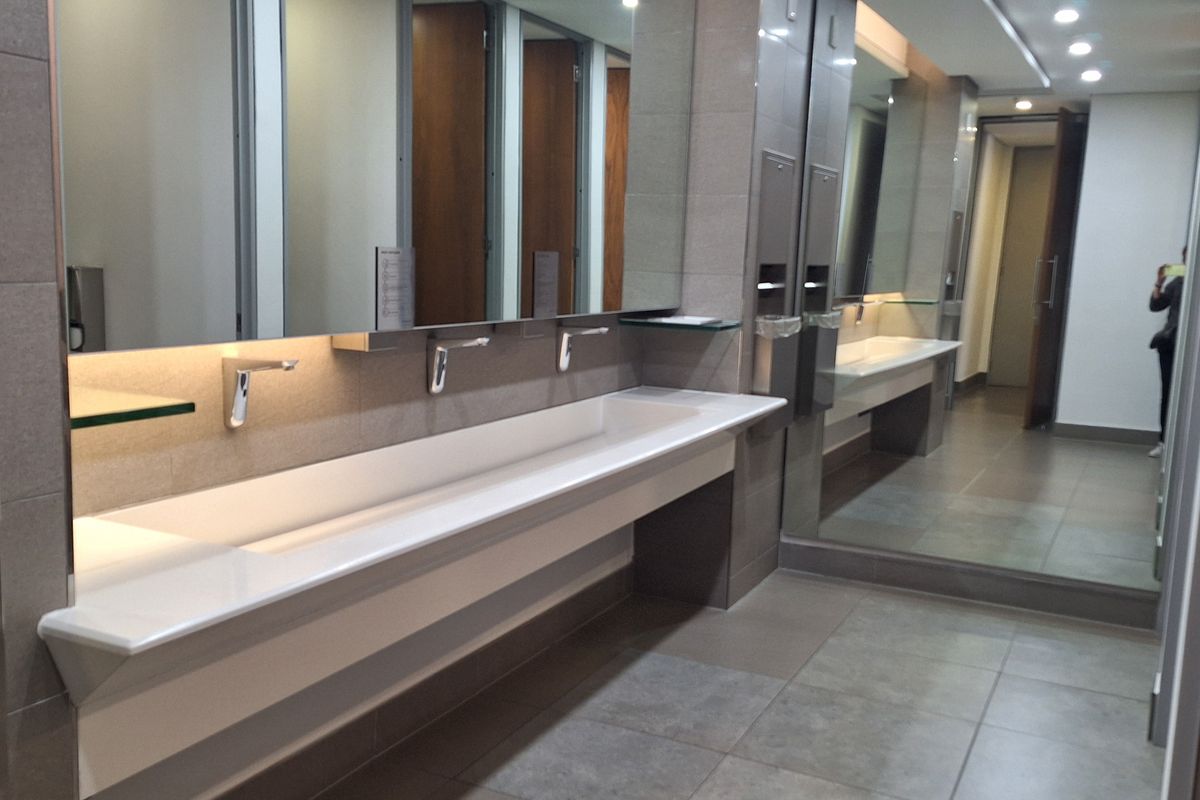 Modern commercial bathroom featuring a long trough sink with wall-mounted faucets, large vanity mirrors, and neutral tiled finishes.