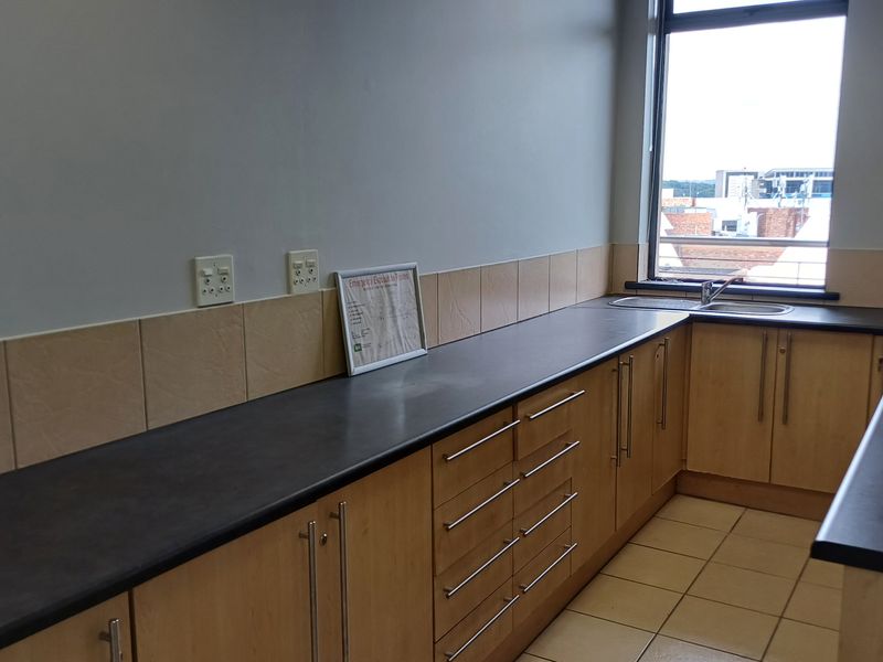 A functional kitchenette featuring light wood cabinetry, dark laminate countertops, a stainless steel sink, and tiled flooring in a professional office setting.