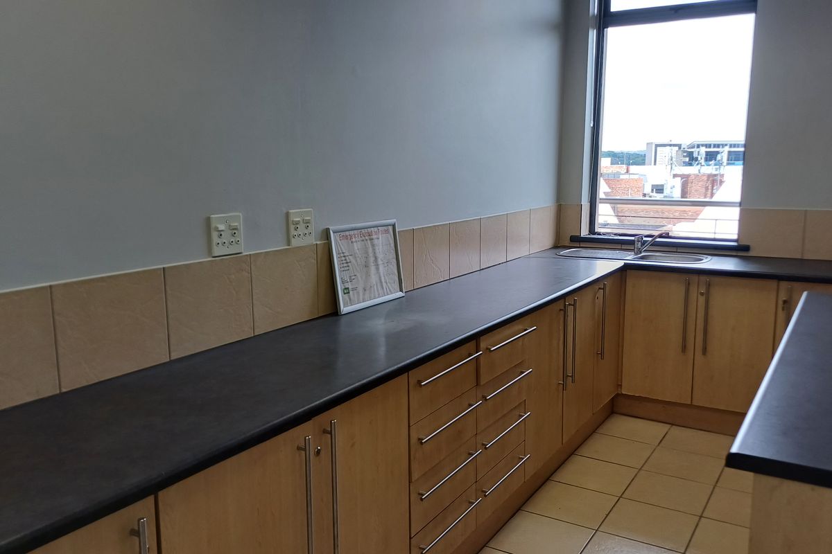 A functional kitchenette featuring light wood cabinetry, dark laminate countertops, a stainless steel sink, and tiled flooring in a professional office setting.