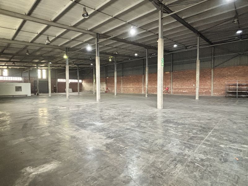 Spacious industrial warehouse featuring high ceilings with exposed steel trusses, concrete flooring, and brick wall construction, suitable for storage or manufacturing operations.