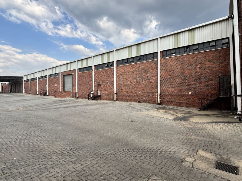 Industrial warehouse exterior featuring a long brick facade, metal cladding, roller shutter access, and a large paved yard area.