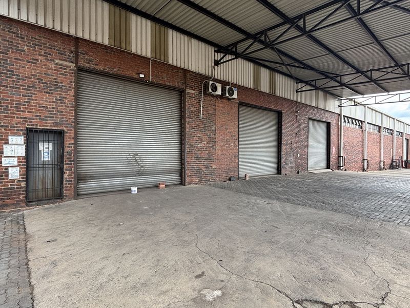 Industrial warehouse exterior featuring multiple roller shutter doors, a covered loading bay, and durable brick construction with paved access area.