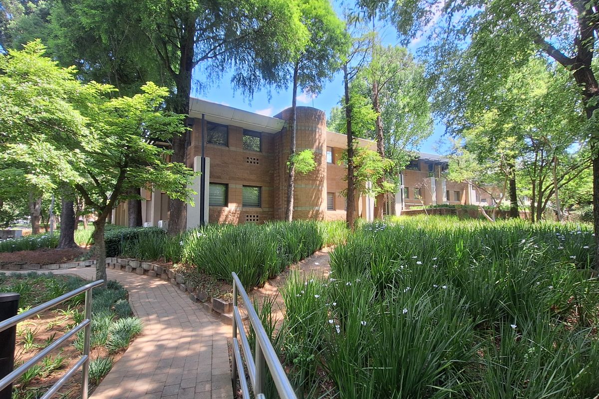 A view of the office building exterior featuring a brick facade, mature trees, and a paved walkway through a landscaped garden.