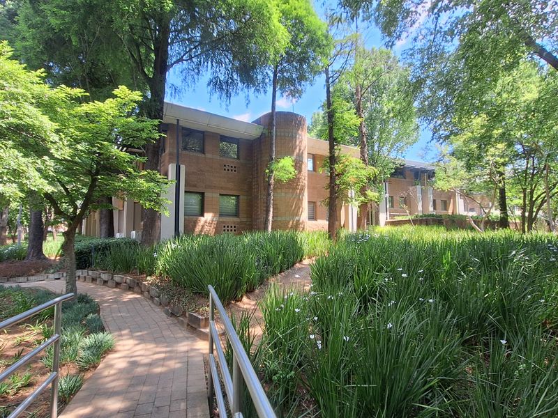 A view of the office building exterior featuring a brick facade, mature trees, and a paved walkway through a landscaped garden.