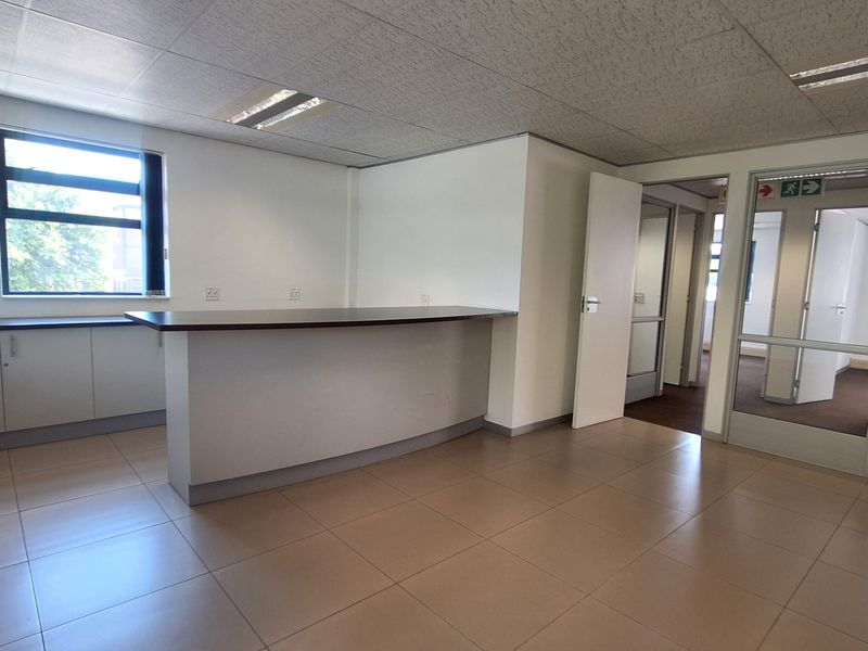 A bright, open-plan office space featuring a built-in kitchenette, tiled flooring, suspended ceilings, and glass partition doors leading to adjacent office areas.