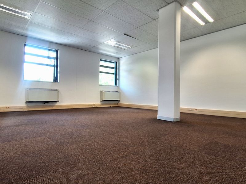 A spacious, empty office area featuring carpet tiles, a suspended ceiling with recessed lighting, and a central structural column.