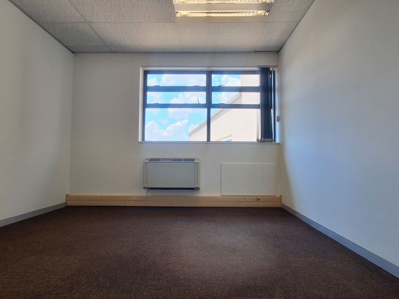 A clean, empty office space featuring carpeted flooring, a suspended ceiling with recessed lighting, and a wall-mounted air conditioning unit.