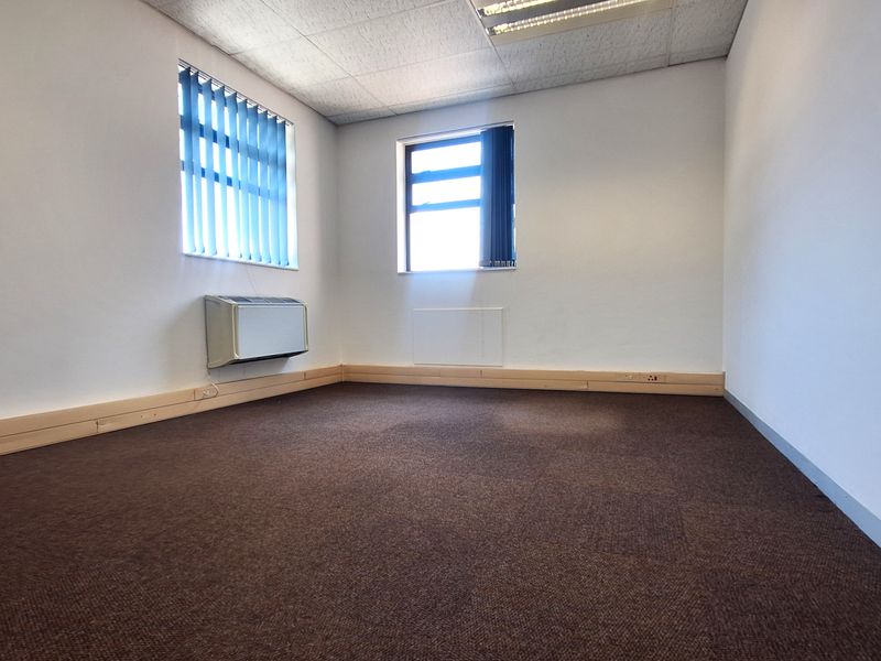 A vacant office space featuring brown carpeting, suspended ceiling tiles, wall-mounted HVAC unit, and vertical blinds on the windows.