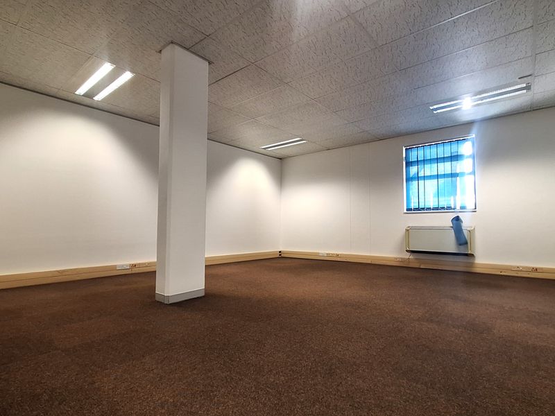 A spacious, empty office area featuring a suspended ceiling, recessed lighting, a structural column, and a window with security bars.