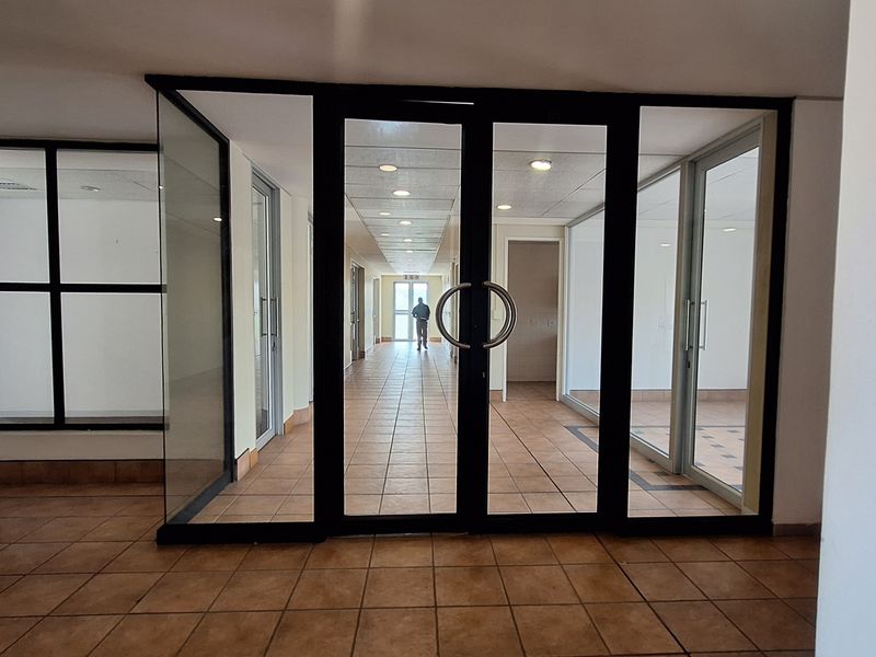A spacious office hallway featuring tiled flooring, recessed ceiling lighting, and glass partition walls with double glass doors leading to the corridor.