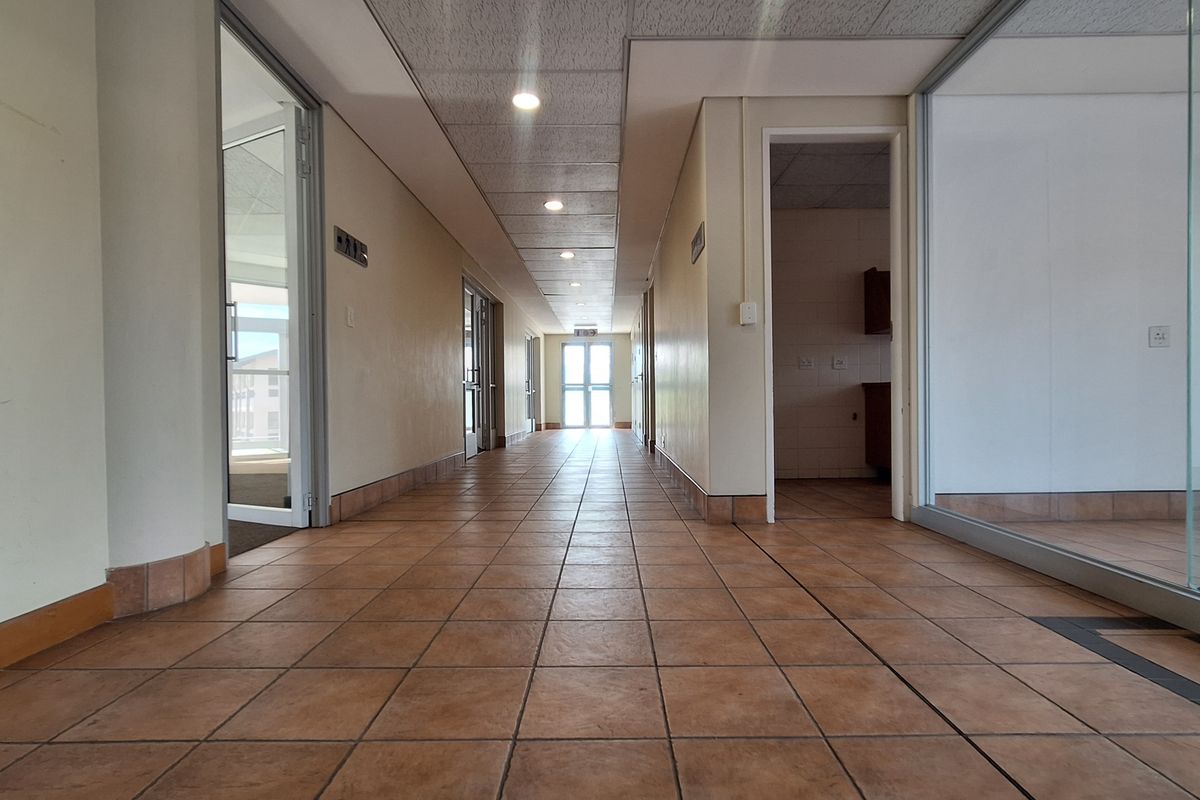 A spacious office hallway featuring tiled flooring, suspended ceilings with recessed lighting, and glass partition walls providing access to various office suites.