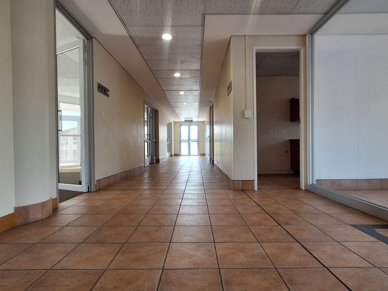 A spacious office hallway featuring tiled flooring, suspended ceilings with recessed lighting, and glass partition walls providing access to various office suites.
