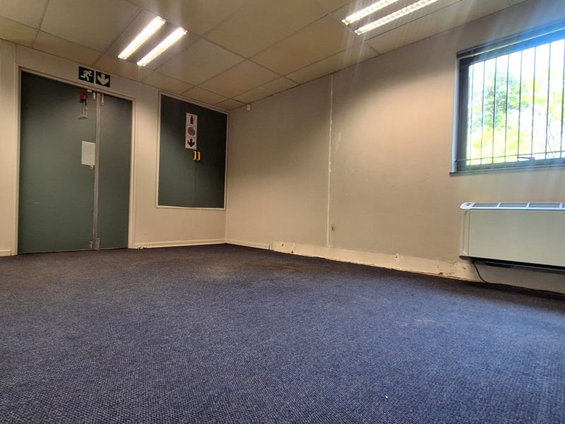 An empty office space featuring carpet flooring, suspended ceiling with recessed lighting, security bars on windows, and a wall-mounted air conditioning unit.