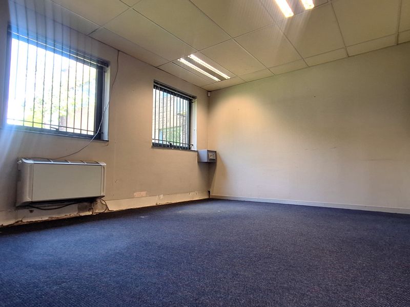 An empty office space featuring carpeted flooring, suspended ceiling with recessed lighting, security bars on windows, and a wall-mounted HVAC unit.