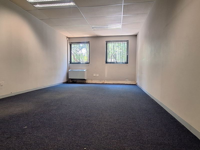 An empty office space featuring a suspended ceiling, recessed lighting, wall-mounted air conditioning, and carpeted flooring with security bars on the windows.
