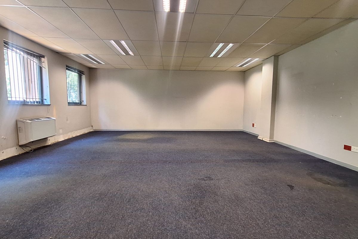 An empty, open-plan office space featuring a suspended ceiling, recessed lighting, carpeted flooring, and a wall-mounted HVAC unit.