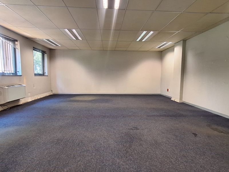 An empty, open-plan office space featuring a suspended ceiling, recessed lighting, carpeted flooring, and a wall-mounted HVAC unit.