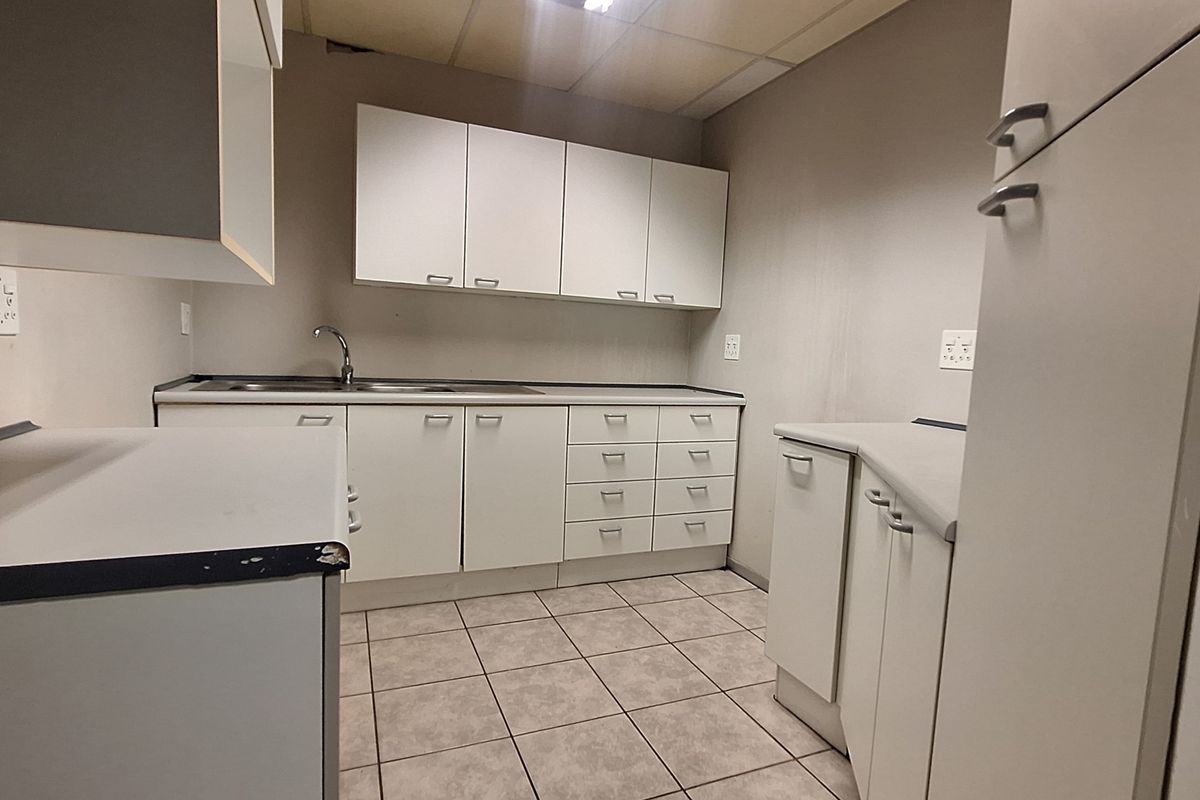 A functional office kitchenette featuring white built-in cabinetry, laminate countertops, a stainless steel sink, and tiled flooring in a neutral color palette.