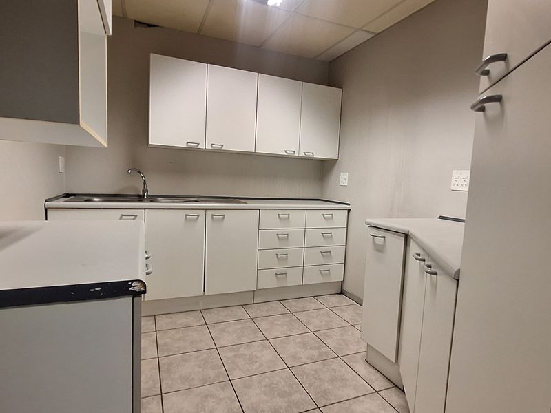 A functional office kitchenette featuring white built-in cabinetry, laminate countertops, a stainless steel sink, and tiled flooring in a neutral color palette.