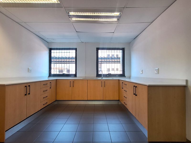 A functional office kitchenette featuring U-shaped built-in cabinetry, laminate countertops, a stainless steel sink, and tiled flooring under a suspended ceiling.