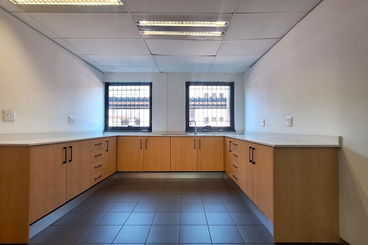 A functional office kitchenette featuring U-shaped built-in cabinetry, laminate countertops, a stainless steel sink, and tiled flooring under a suspended ceiling.