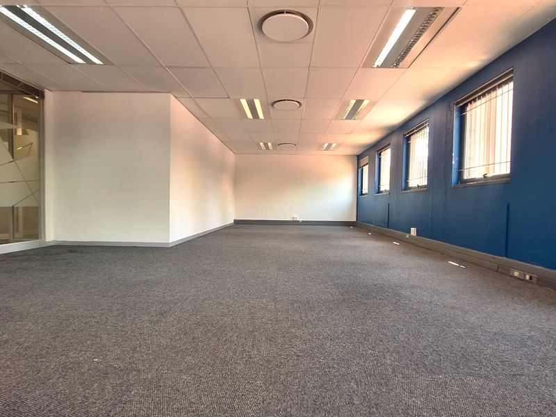 A spacious, empty office area featuring carpeted flooring, a suspended ceiling with recessed lighting, and large windows with security bars.