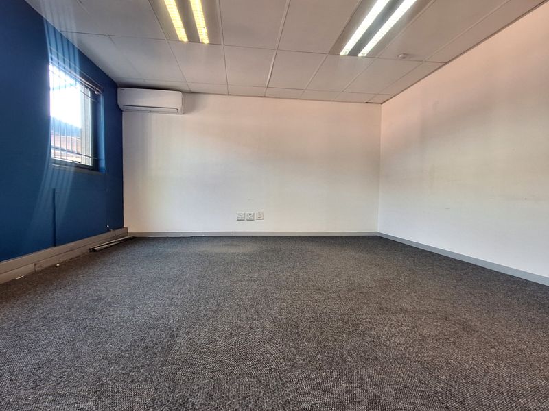 An empty office space featuring grey carpeting, a suspended ceiling with recessed lighting, and a wall-mounted air conditioning unit.