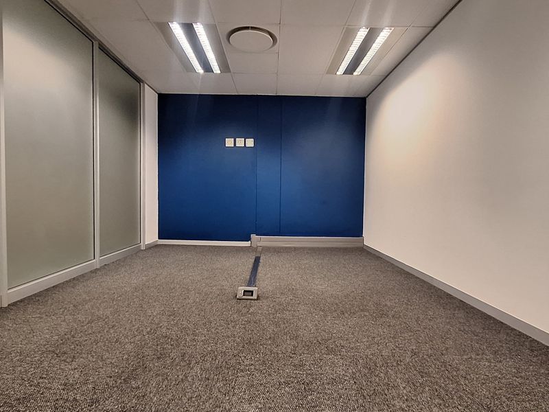 A clean, empty office space featuring carpeted flooring, a suspended ceiling with recessed lighting, and a modern glass partition wall.