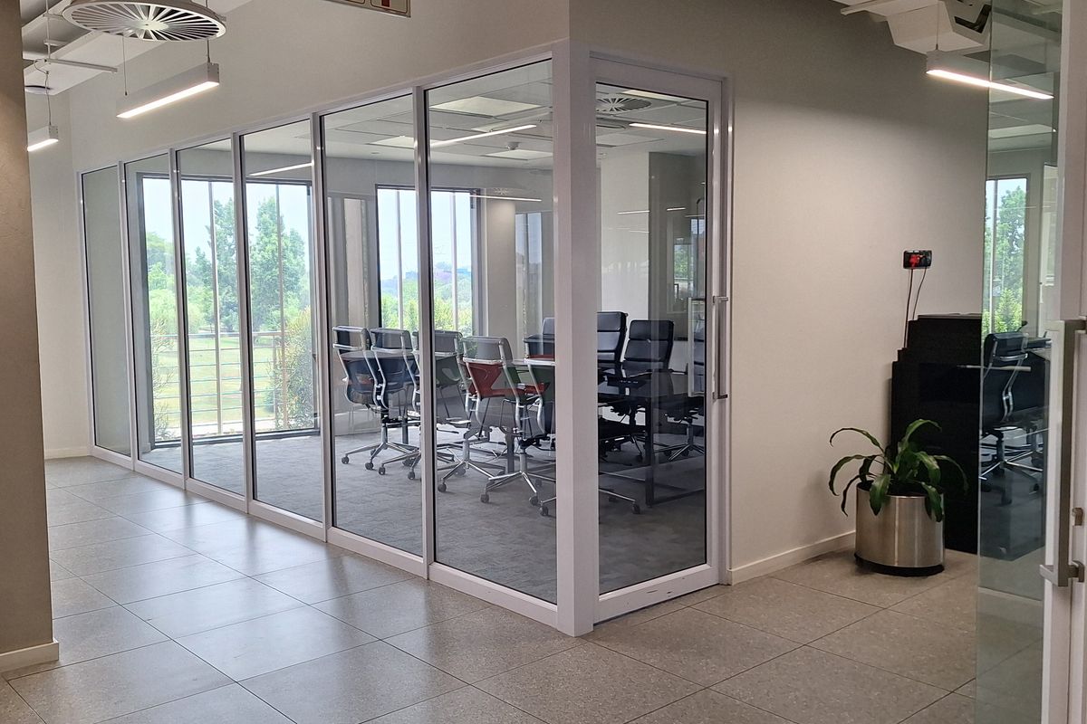 Modern office space featuring glass-partitioned meeting rooms, tiled flooring, and contemporary exposed ceiling infrastructure with integrated lighting.
