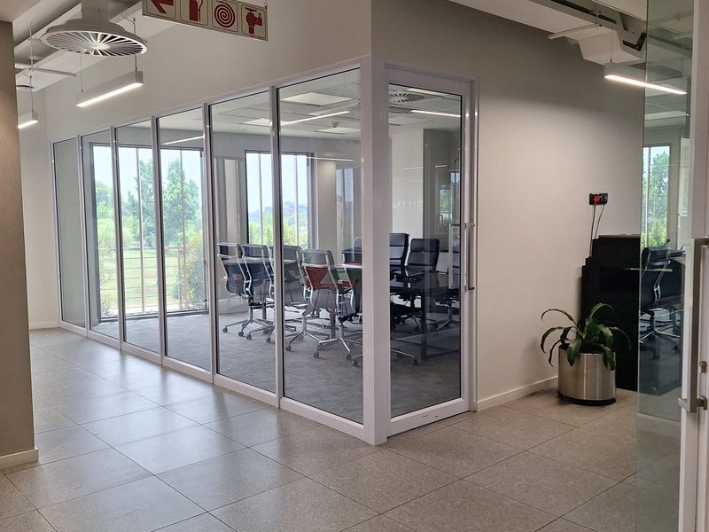 Modern office space featuring glass-partitioned meeting rooms, tiled flooring, and contemporary exposed ceiling infrastructure with integrated lighting.