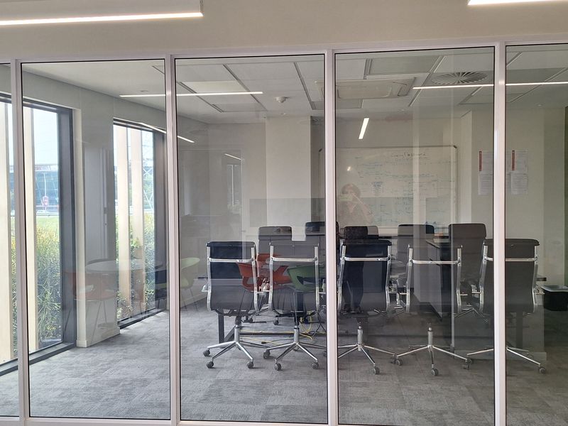 A modern office meeting room enclosed by floor-to-ceiling glass partitions, featuring carpeted flooring, integrated lighting, and a wall-mounted whiteboard.