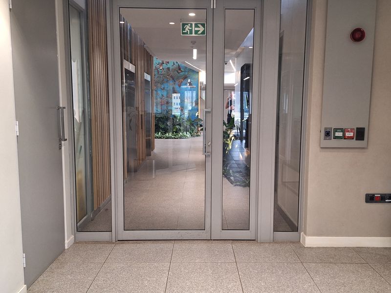 Modern office building entrance featuring glass double doors, emergency exit signage, fire safety equipment, and contemporary timber slat wall detailing.