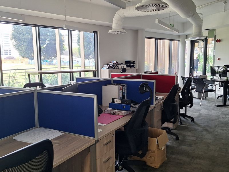 Modern open-plan office space featuring large windows, exposed industrial-style ductwork, and integrated workstation partitions with carpeted flooring.