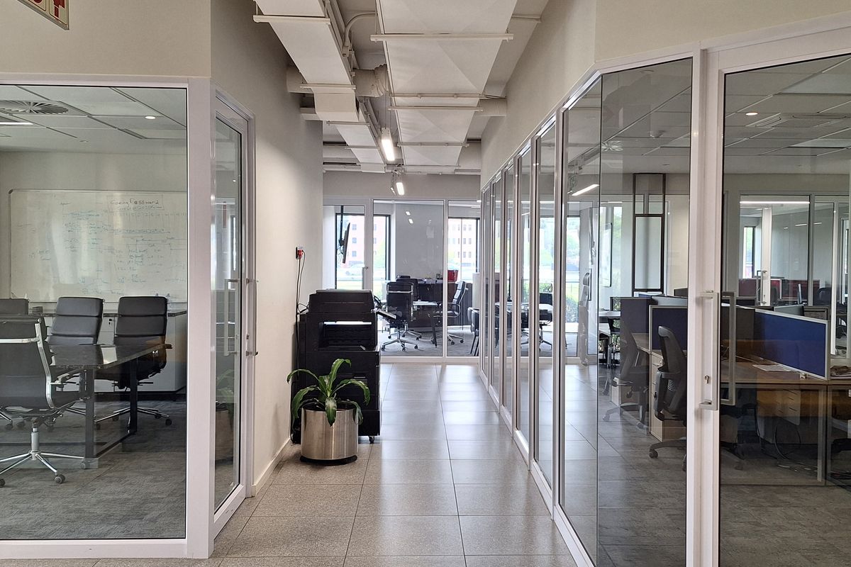 A modern office hallway featuring glass-partitioned meeting rooms and workspaces, exposed ceiling ducting, and clean tiled flooring.