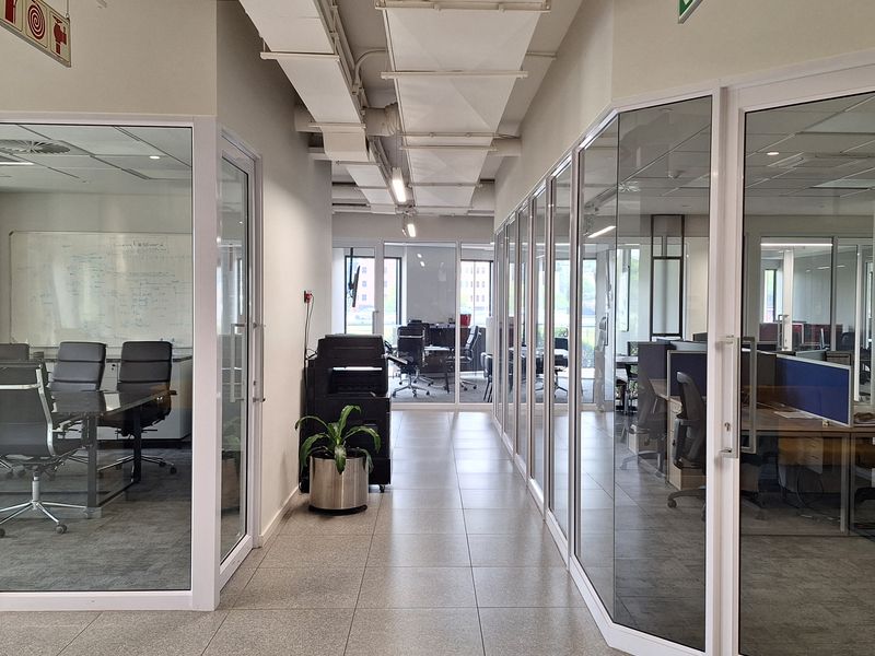 A modern office hallway featuring glass-partitioned meeting rooms and workspaces, exposed ceiling ducting, and clean tiled flooring.