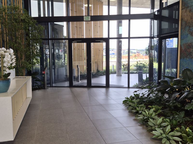 Modern office building entrance featuring a sleek reception desk, floor-to-ceiling glass facade, and high-quality tiled flooring with integrated indoor landscaping.