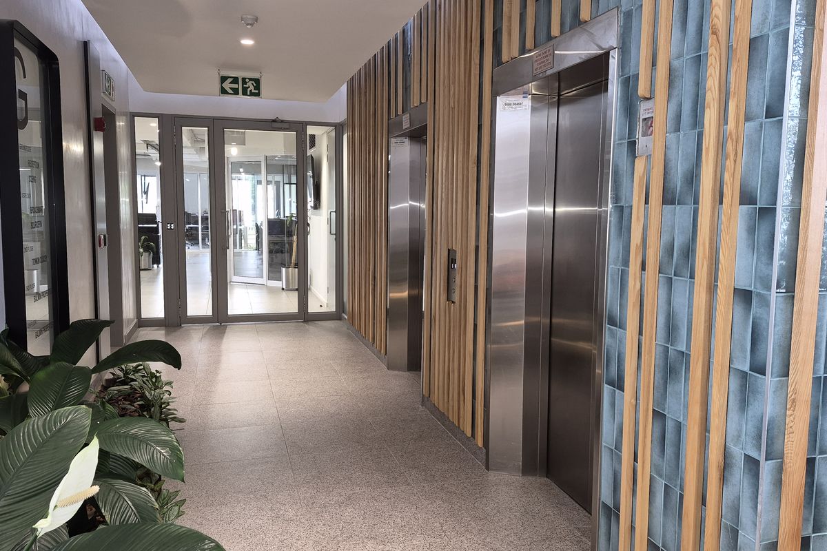 Modern office building lobby featuring stainless steel elevators, decorative timber slat wall paneling, blue feature tiling, and glass-panelled office entrance doors.