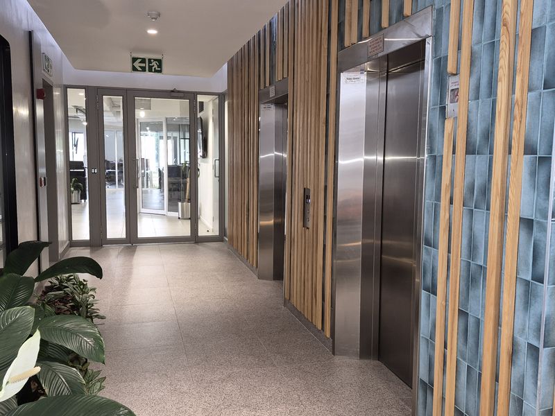 Modern office building lobby featuring stainless steel elevators, decorative timber slat wall paneling, blue feature tiling, and glass-panelled office entrance doors.