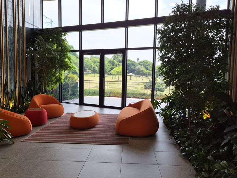 Modern office lobby featuring floor-to-ceiling glass walls, tiled flooring, and integrated indoor greenery with views of the surrounding landscape.