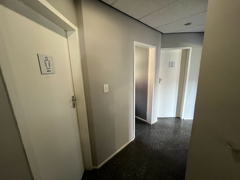 A clean office hallway featuring tiled flooring, a drop ceiling with recessed lighting, and clearly marked restroom access doors.