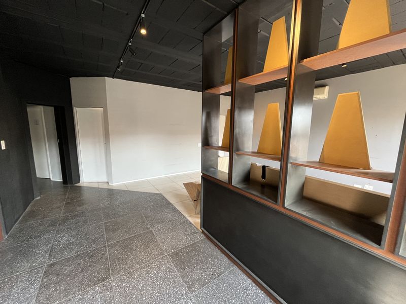 A modern, open-plan office space featuring dark tiled flooring, a built-in shelving partition, and an exposed ceiling with track lighting.