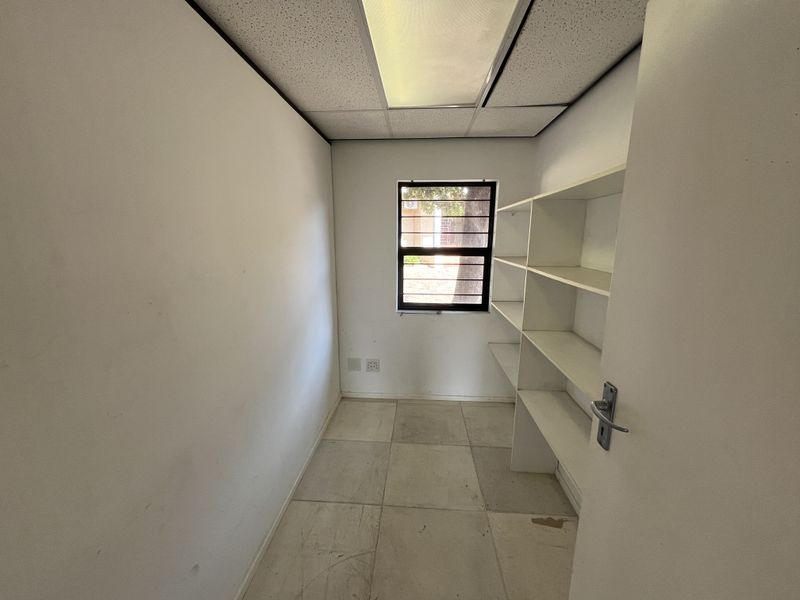 A small, empty office space featuring built-in shelving, tiled flooring, and a drop ceiling with integrated lighting.