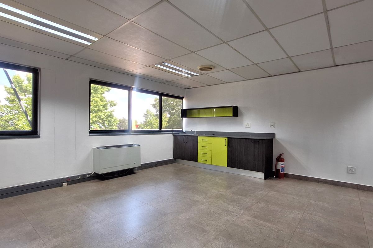 A bright, open-plan office space featuring a built-in kitchenette, tiled flooring, suspended ceiling, and large windows providing ample natural light.