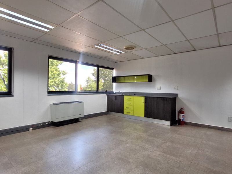 A bright, open-plan office space featuring a built-in kitchenette, tiled flooring, suspended ceiling, and large windows providing ample natural light.